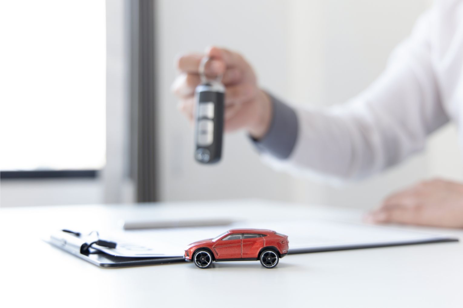 Mão de pessoa branca, vestindo camisa de manga longa branca. A mão direita está segurando a chave de um carro enquanto a mão esquerda repousa sobre um contrato de seguro de automóvel. Um carro de brinquedo vermelho fica em foco, enquanto o resto da imagem fica desfocada.
