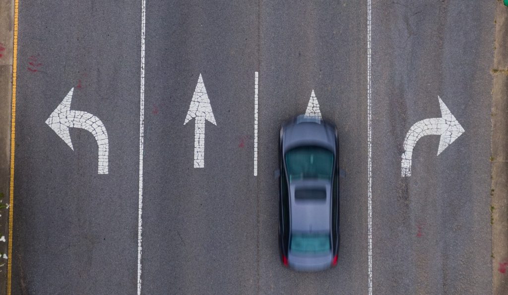 Vista aérea de carro cinza parado na rua em que constam sinalizações em branco a direita e a esquerda.