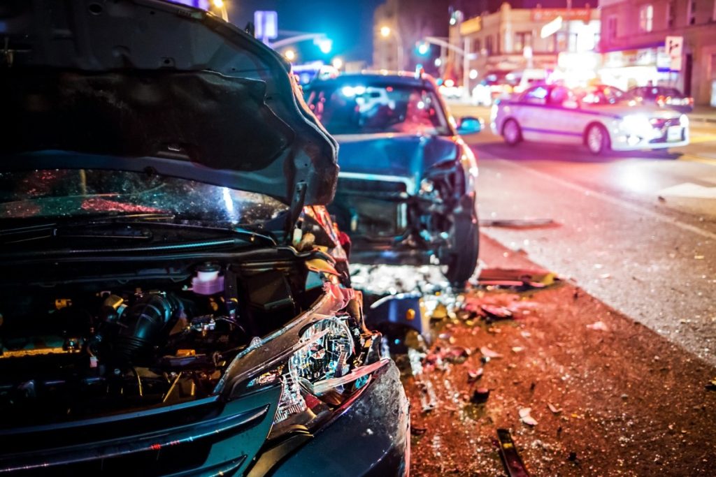 Seguro auto é essencial em casos de acidente, como mostra essa foto. A imagem é de dois carros envolvidos em um acidente de noite. No fundo, há um carro de polícia com as luzes acessas.