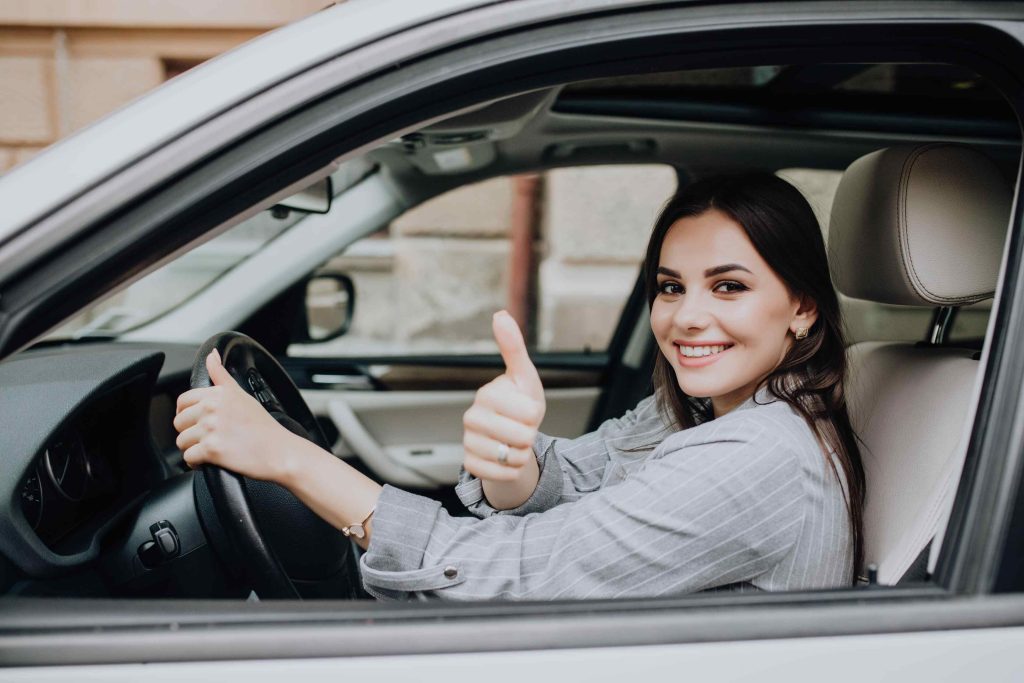 Mulher de cabelo castanho e vestindo uma camisa cinza fazendo gesto de joinha com a mão direita, enquanto posiciona a mão esquerda no volante de um carro cinza.