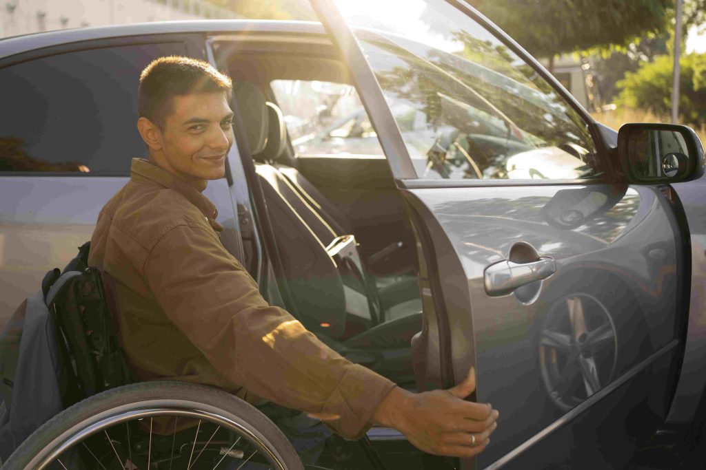Homem sorridente em uma cadeira de rodas, sorrindo. Está abrindo a porta de um carro cinza.