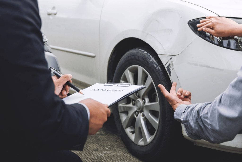 A franquia de seguro auto é importante, conforme demonstra essa imagem. Duas pessoas estão avaliando um carro branco que foi danificado. Um deles segura uma prancheta.