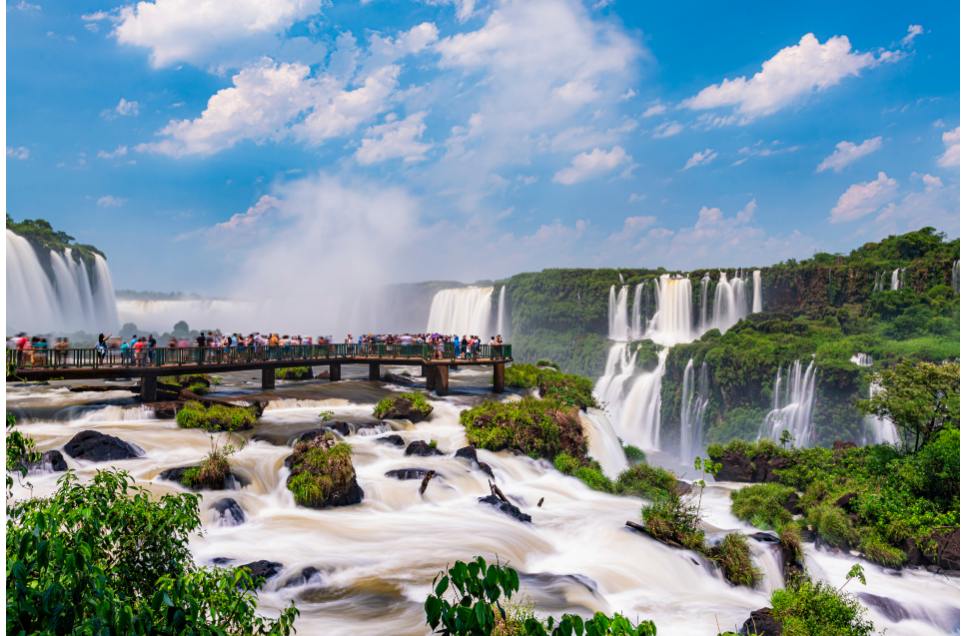 Turistas caminhando em uma passarela sobre as águas das Cataratas do Iguaçu, situadas no Paraná. O céu azul e com poucas nuvens reflete-se na superfície, enquanto a vegetação circunda a paisagem, proporcionando um ambiente naturalmente deslumbrante. Cote agora o seu seguro auto no Paraná!