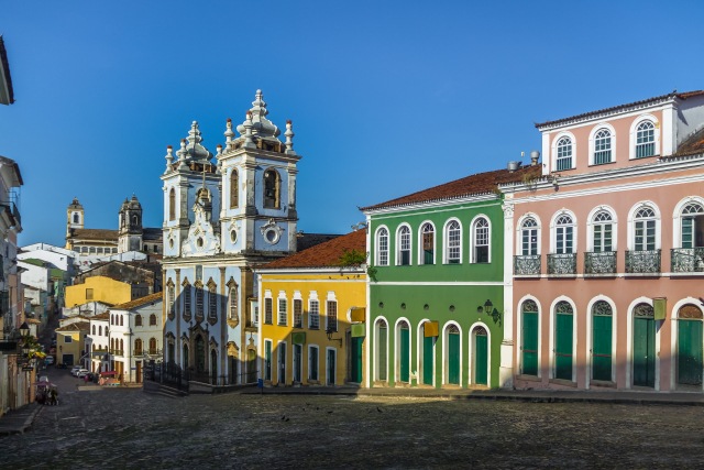 Parte da cidade histórica de Salvador. Uma rua paralelepípedo com prédios em estilo colonial, pintados em cores vibrantes. O céu é um azul intenso. Contrate agora seu seguro auto em Salvador!