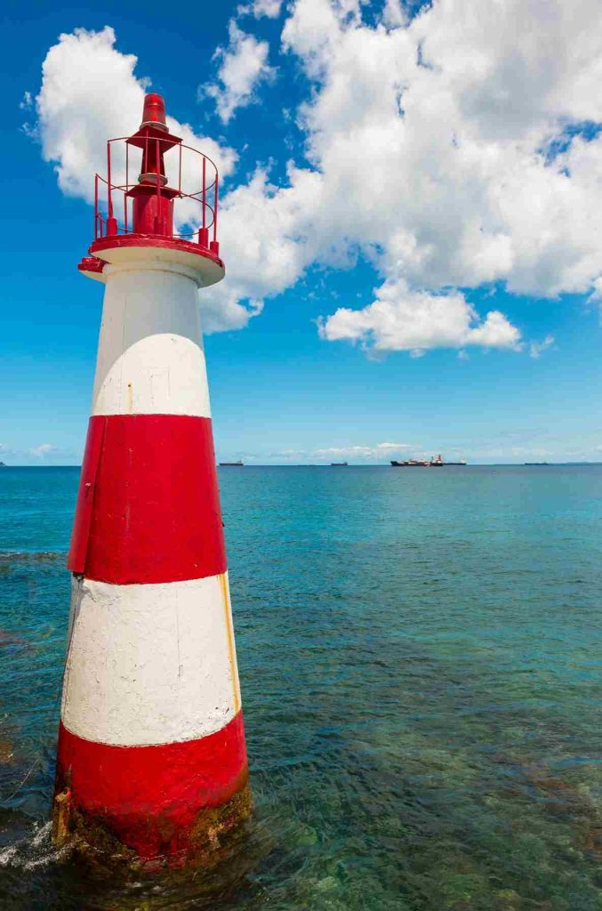 Na praia de Salvador, Bahia, há um farol branco e vermelho. No horizonte, navios são visíveis. Ao fundo, montanhas. A água do mar é azul. O céu tem nuvens brancas e esparsas. Para este passeio contrate hoje seu seguro auto na Bahia