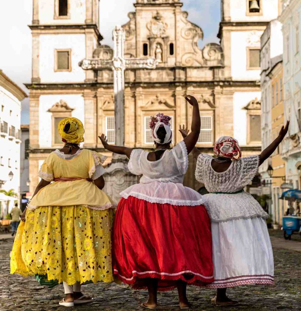 Na Bahia, há festas culturais com mulheres usando vestimentas típicas, saias rodadas e coloridas. Montanhas ao fundo. Colares, pulseiras e turbantes representam a cultura africana